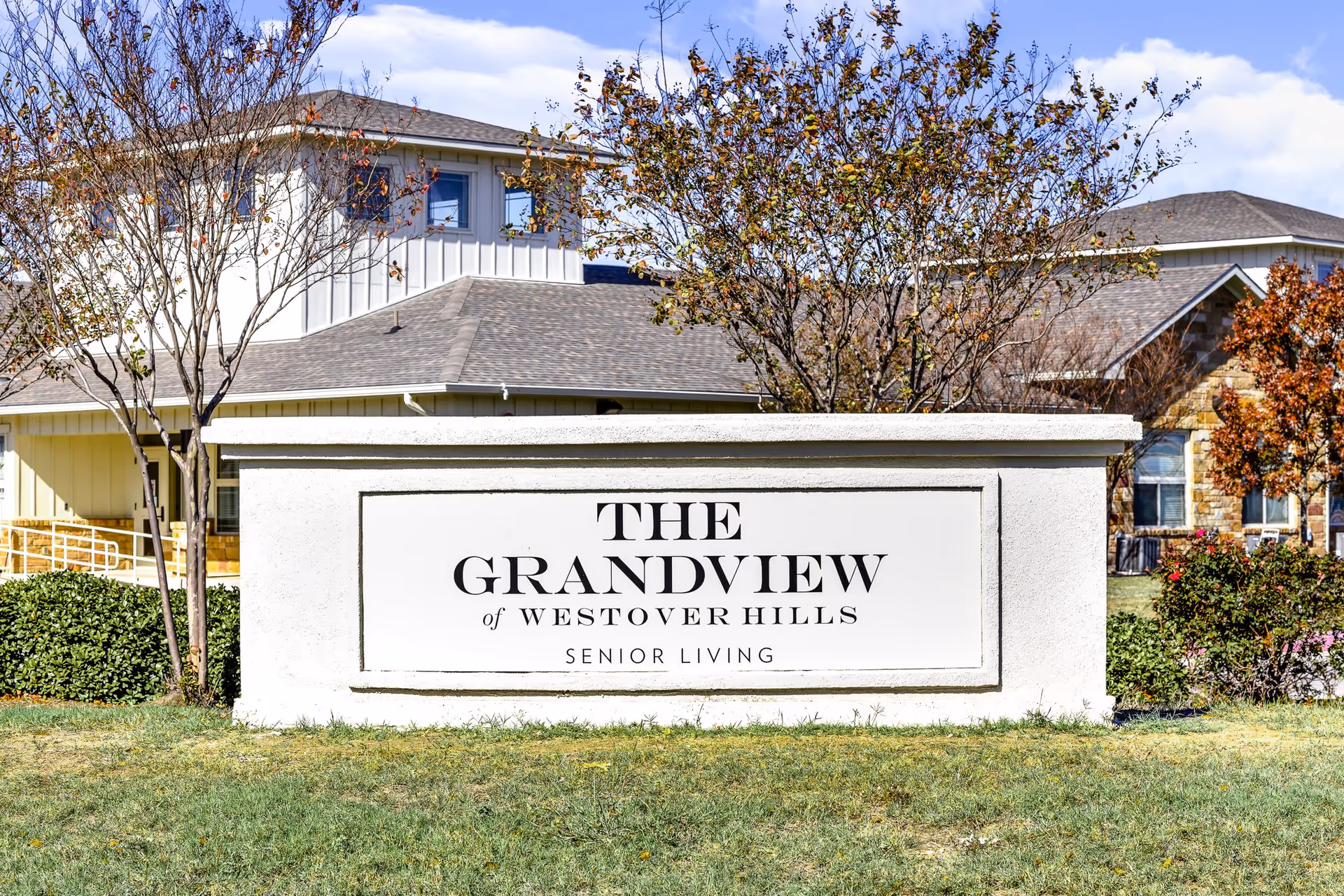 Outdoor view of a large white sign with black text that reads 'The Grandview of Westover Hills Senior Living' in front of a senior living facility building with trees and bushes around.