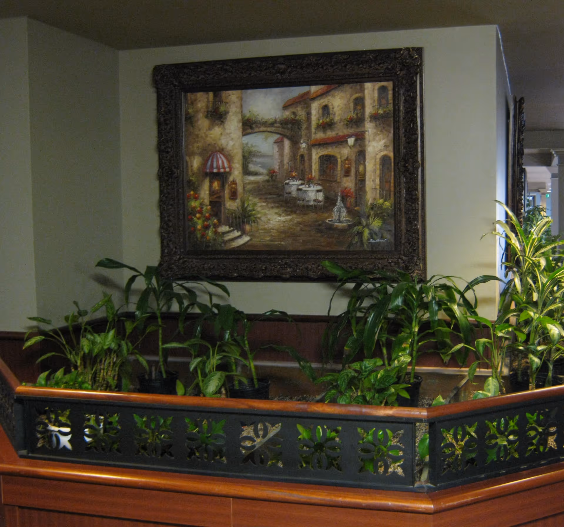 Indoor corner planter with various green plants surrounded by a decorative black metal railing with wooden trim. Above the planter, a framed painting depicts a quaint European street scene with stone buildings, outdoor tables, and flowers.