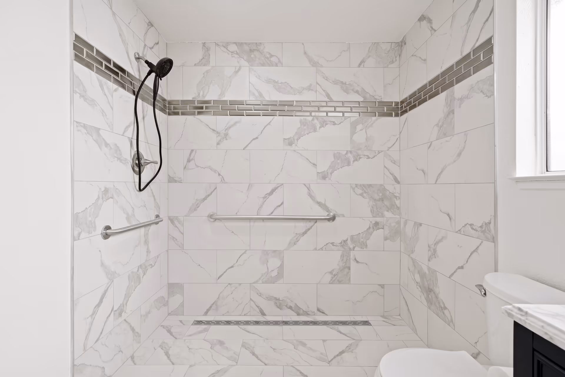 A modern bathroom shower area with white and gray marble-patterned tiles covering the walls and floor. The shower features a black handheld showerhead attached to the wall, two horizontal stainless steel grab bars, and a linear drain on the floor. Part of a white toilet and a marble countertop with a black cabinet are visible on the right side, along with a frosted window.