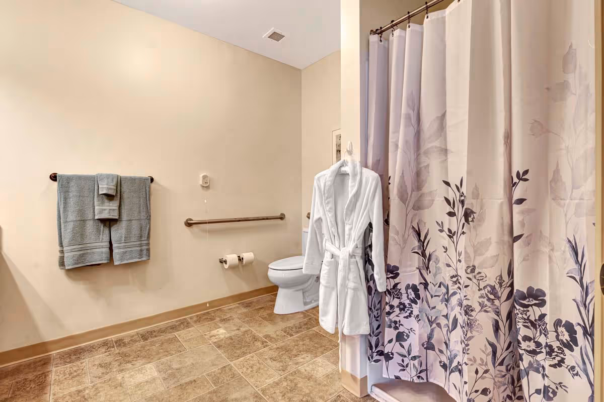 A bathroom with beige walls and tiled floor featuring a toilet, a shower with a floral-patterned curtain, a white bathrobe hanging on the shower wall, a towel rack with two gray towels, and a grab bar near the toilet.