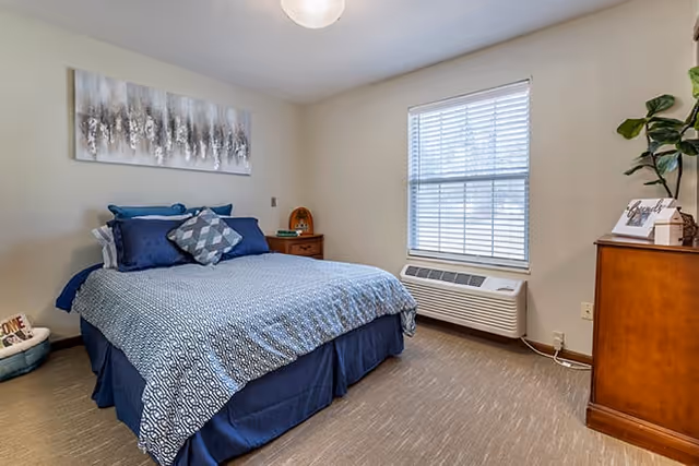 Bright bedroom with a bed dressed in blue linens, nightstands, a window with blinds, and a wooden dresser.