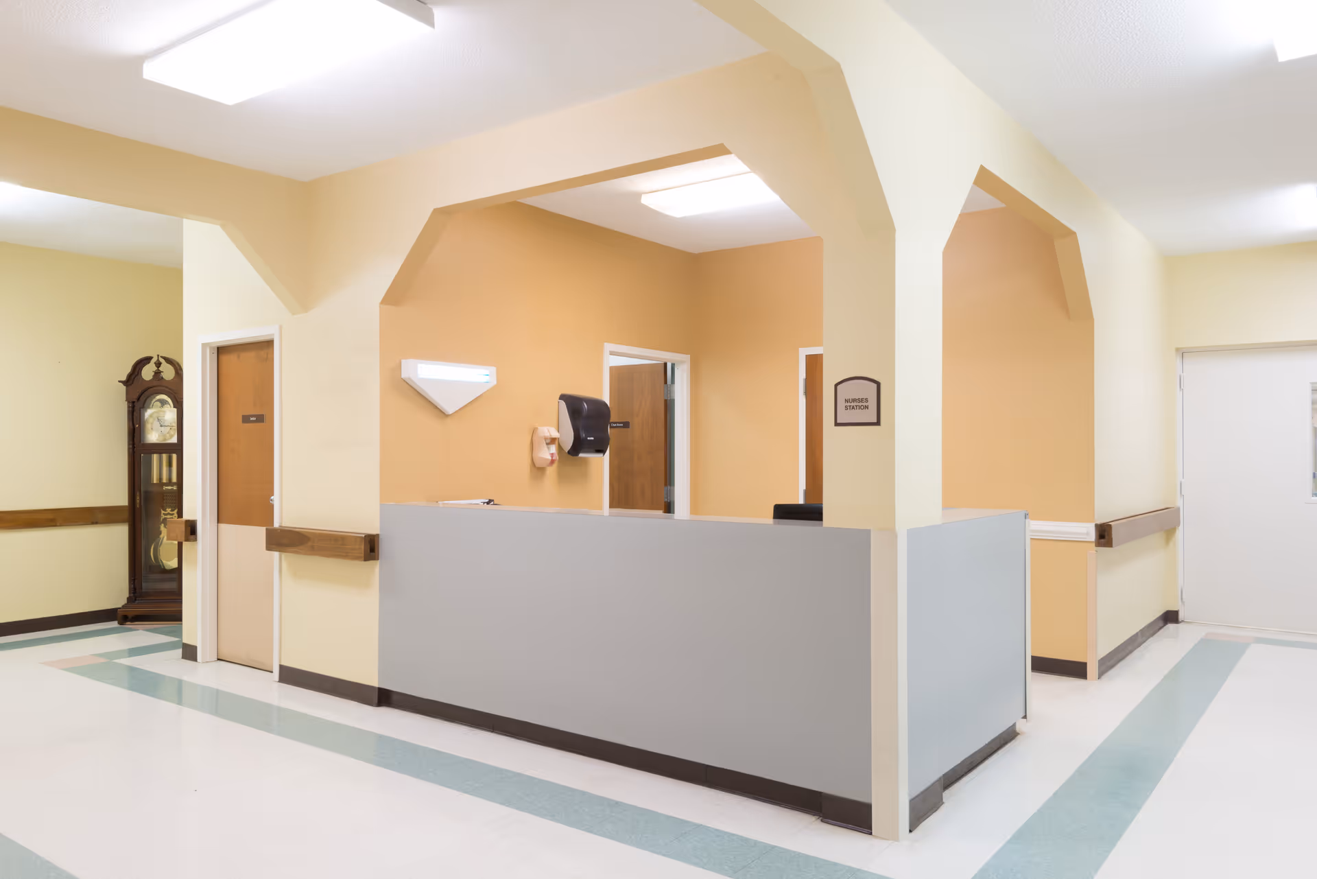 Interior view of a nurse's station in a senior care facility with beige and light yellow walls, a gray counter, wooden doors, a wall-mounted paper towel dispenser, and a grandfather clock in the hallway.