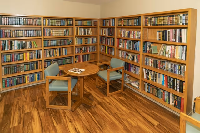 A cozy reading nook with wooden bookshelves filled with books lining two walls. In the center, there is a small round wooden table with two wooden chairs that have light blue cushions. The floor is made of polished wood, and the walls are painted a light color.