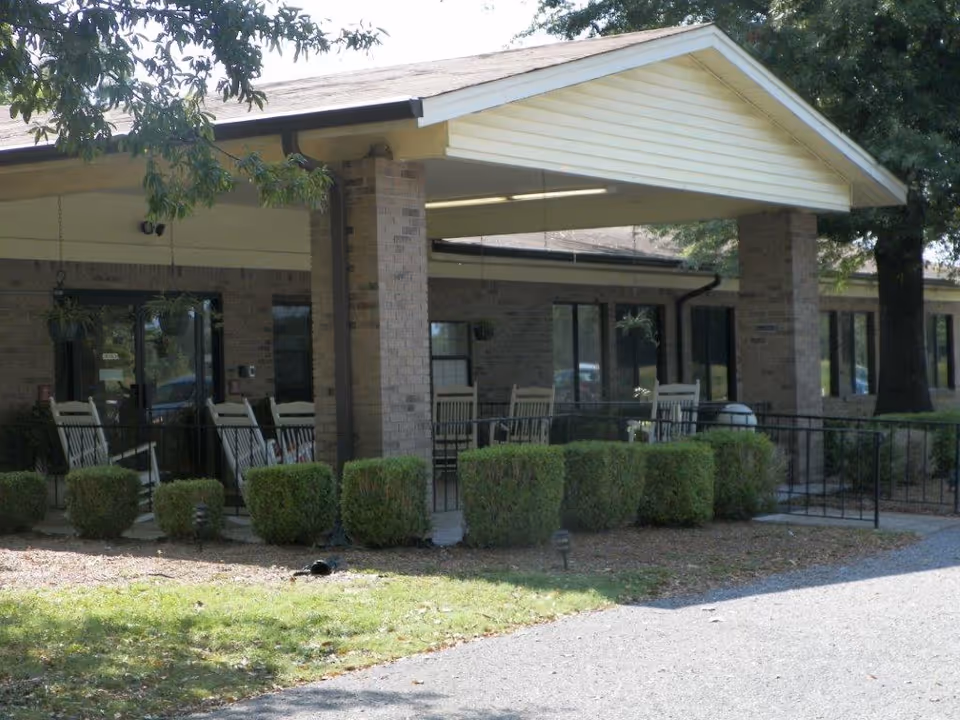 Exterior view of Ripley Healthcare & Rehabilitation Center showing a covered entrance with brick pillars, several rocking chairs on the porch, trimmed bushes, and a paved driveway.