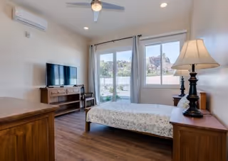 A bright bedroom with a single bed covered in a floral bedspread, wooden furniture including a dresser and nightstand with a lamp, a flat-screen TV on a wooden stand, a ceiling fan, and large windows with curtains showing an outdoor view of rocky hills.