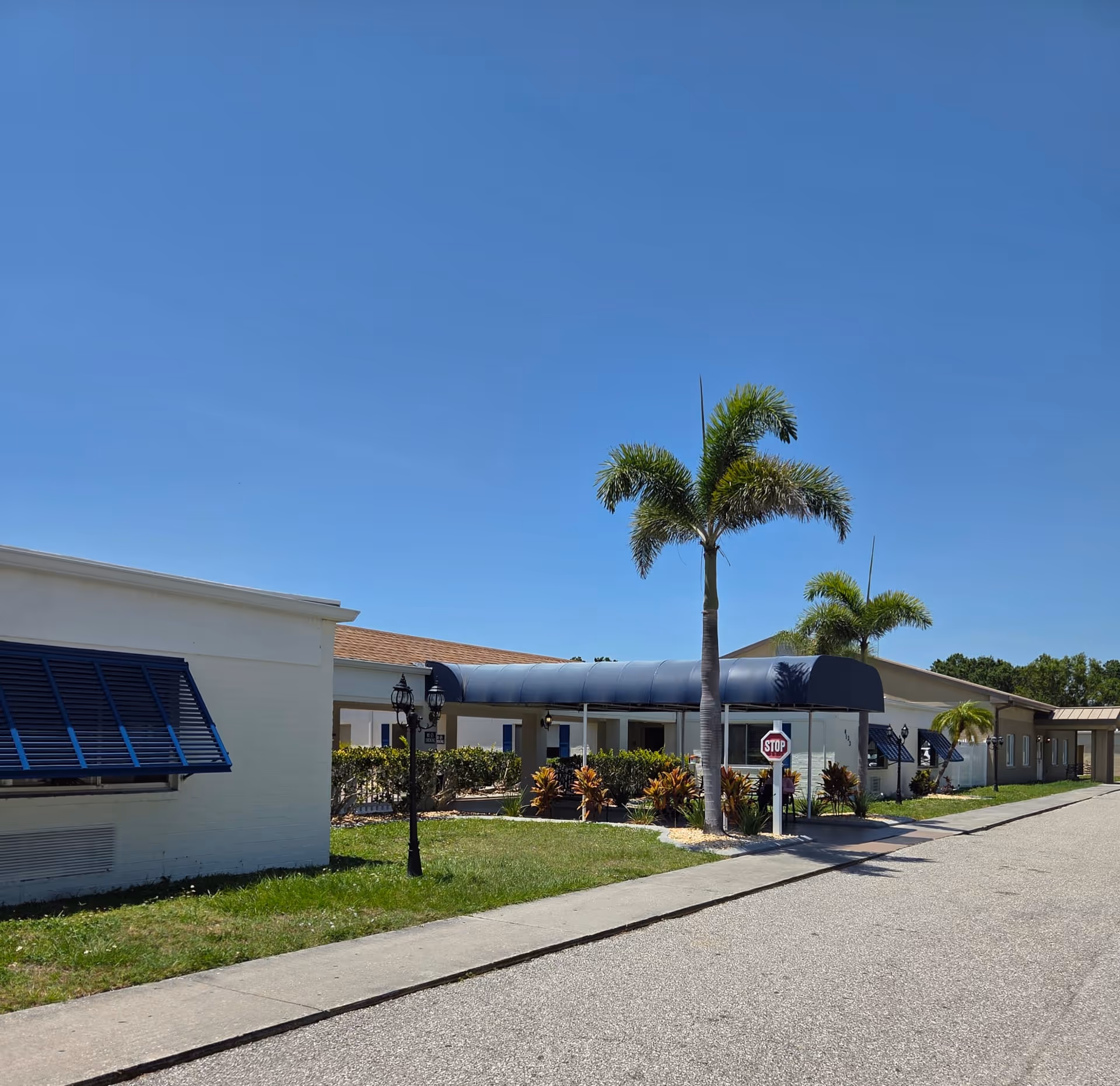 Exterior view of Charlotte Bay Rehabilitation & Care Center on a clear sunny day, showing a single-story building with a blue awning over the entrance, palm trees, a stop sign, and a paved driveway.