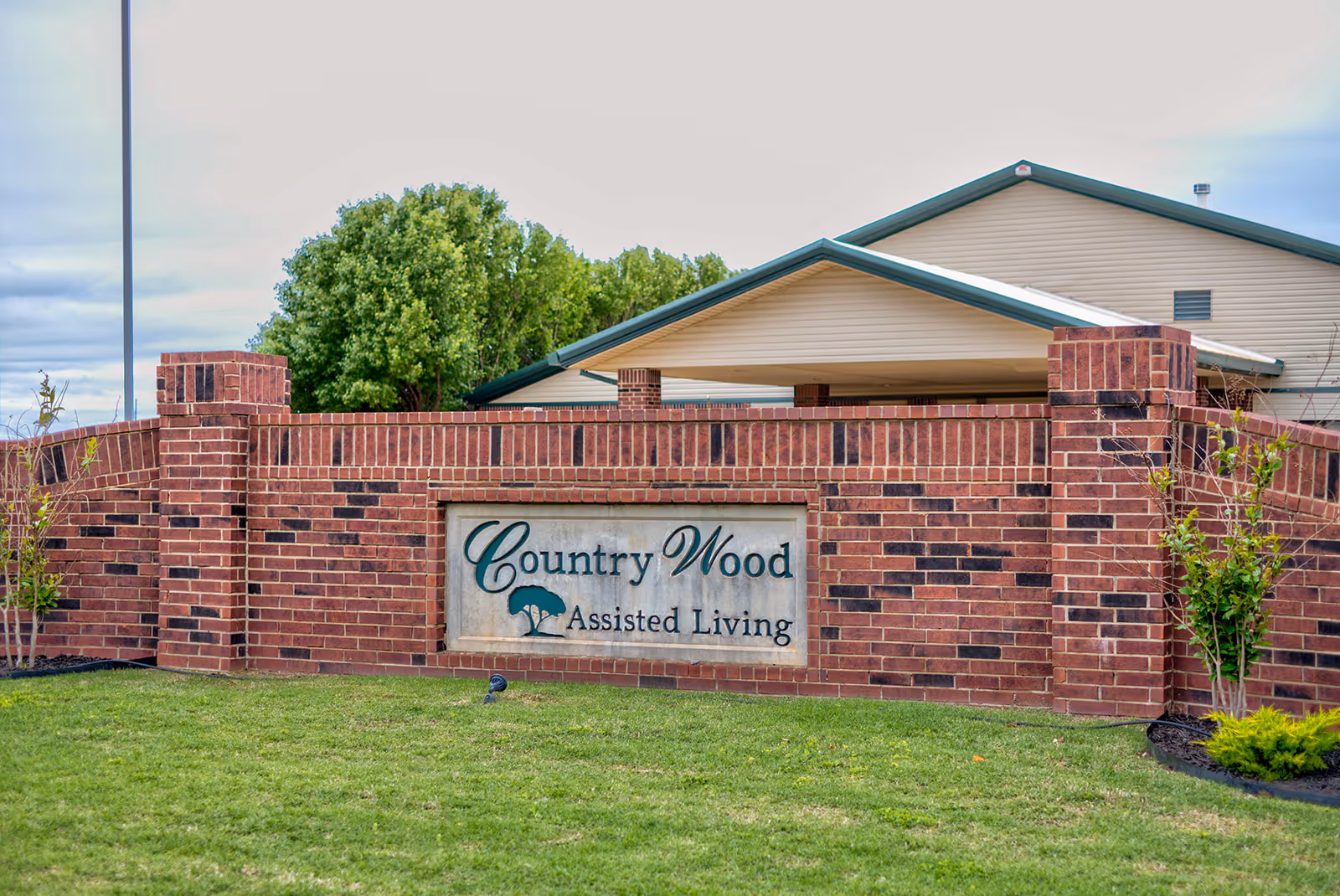 Brick entrance sign for Country Wood Assisted Living with a building and green trees in the background under a cloudy sky.