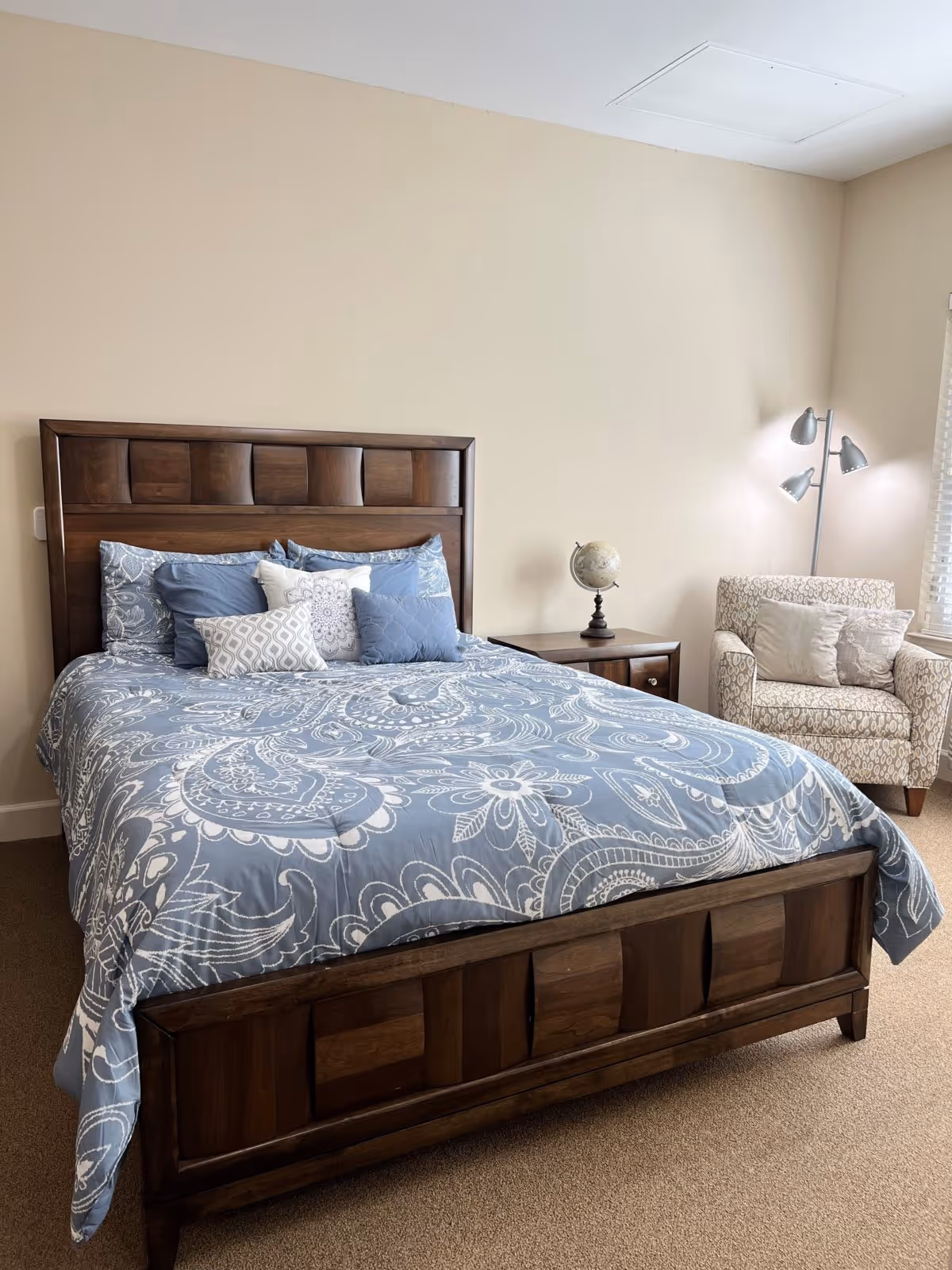 A bedroom with a wooden bed frame featuring a blue and white patterned comforter and multiple pillows. Next to the bed is a wooden nightstand with a small globe on it. In the corner of the room is a patterned armchair with two cushions and a three-headed floor lamp. The walls are beige and there is a window with blinds partially visible.