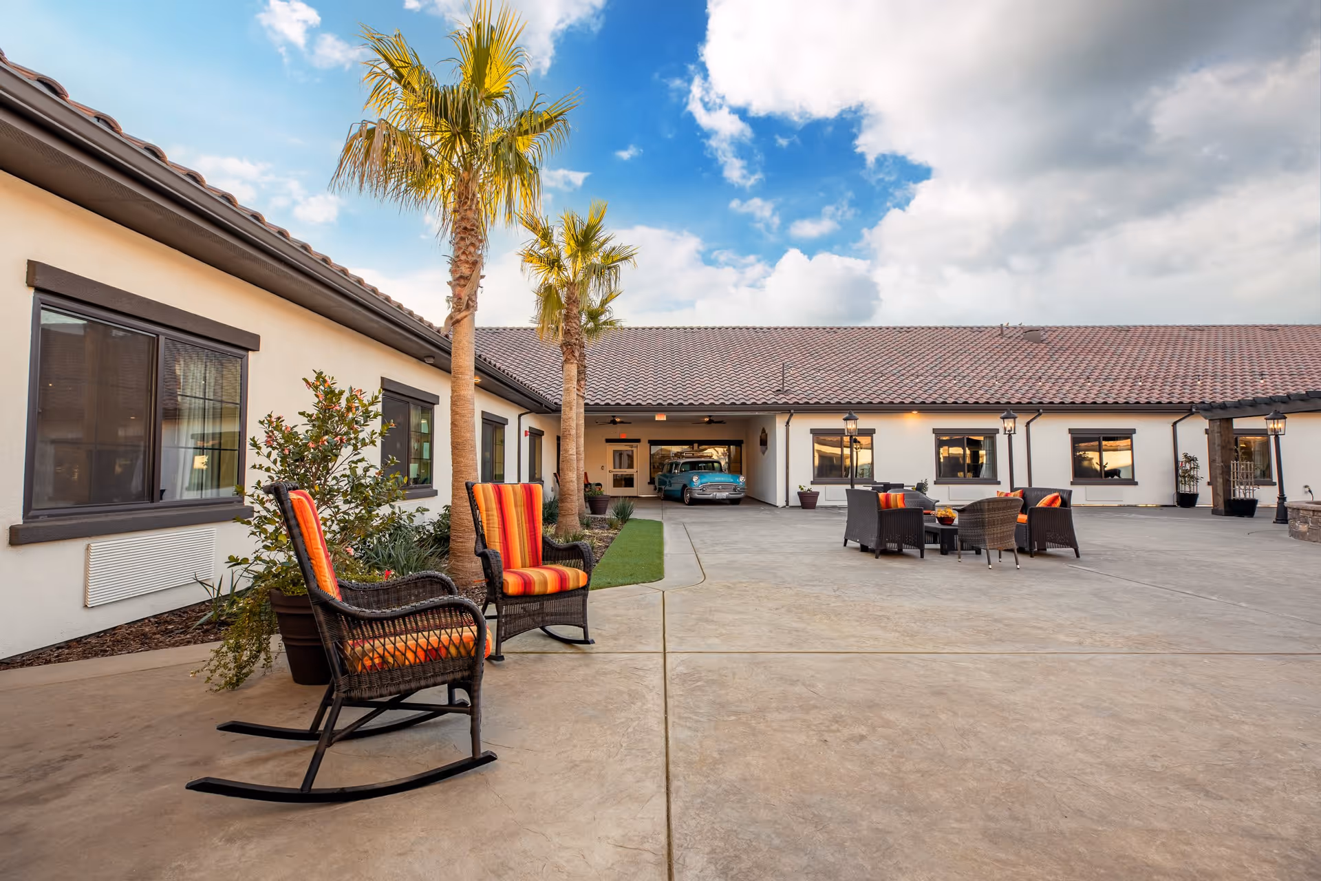 Outdoor courtyard area of a senior living facility with two wicker rocking chairs with orange and red striped cushions, palm trees, potted plants, and a seating area with wicker chairs and a table. The building has a tiled roof and cream-colored walls with dark trim around the windows. A vintage blue car is parked in a garage in the background under a partly cloudy sky.