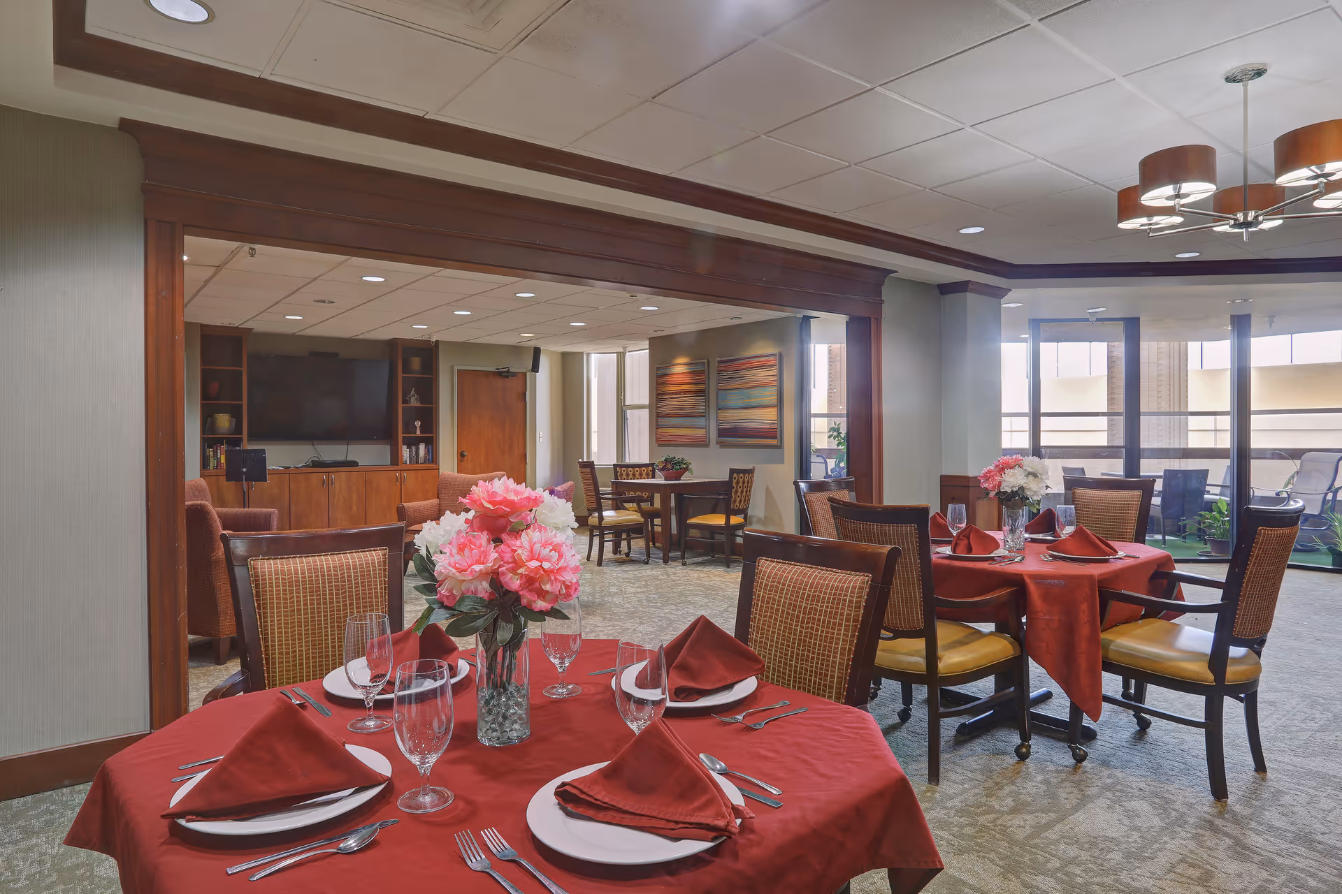 A dining area in a senior living facility with round tables covered in red tablecloths, each set with plates, silverware, glasses, and red napkins. Pink and white flower centerpieces adorn the tables. The room has large windows letting in natural light, and a seating area with a TV and wooden cabinets is visible in the background.