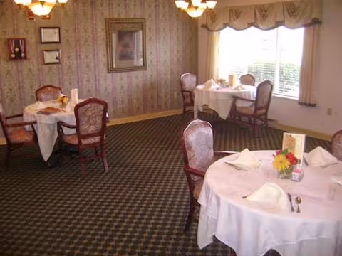 Small dining room with round tables dressed in white tablecloths and upholstered chairs by a curtained window.
