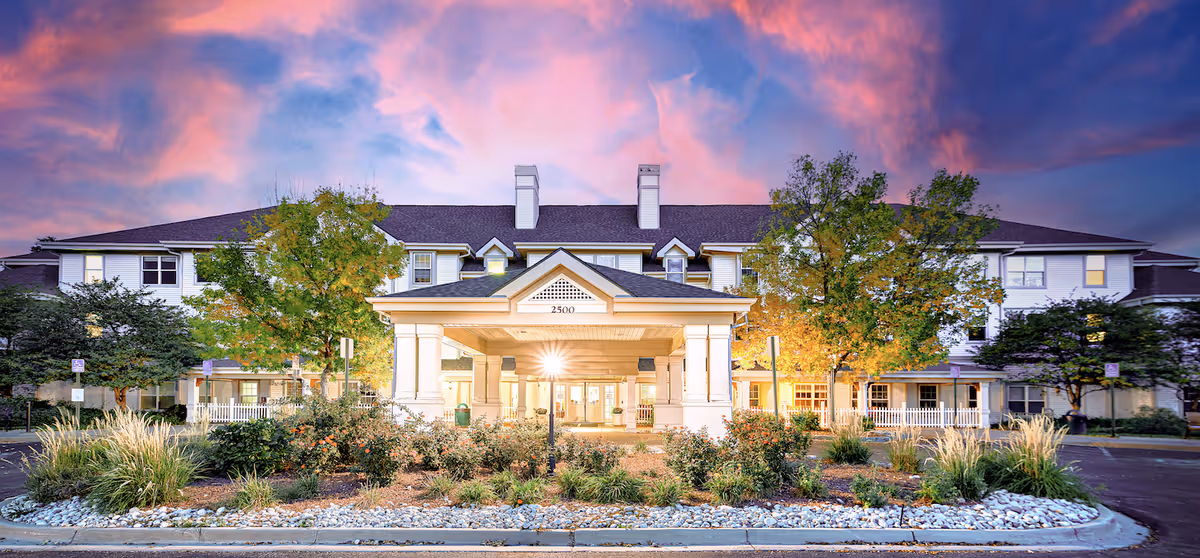 Front exterior view of a large senior living facility building named Woodland Estates Senior Living, with a covered entrance, landscaped garden with bushes and trees, and a dramatic pink and purple sunset sky in the background.