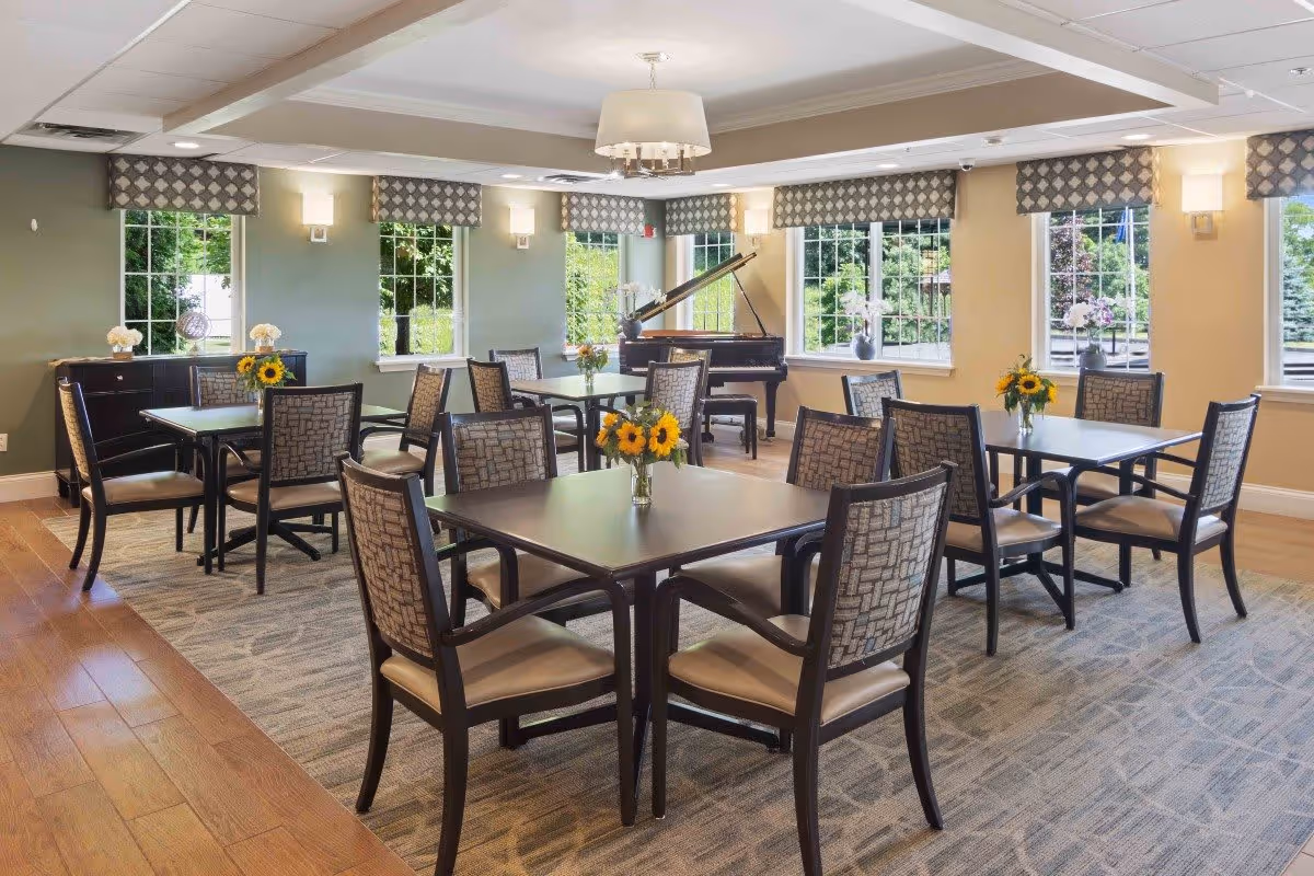 A bright and spacious dining room with multiple square tables, each surrounded by four cushioned chairs. The tables have small vases with sunflowers. Large windows with patterned valances allow natural light to fill the room. A grand piano is positioned near the windows in the background. The room features a mix of wood flooring and carpet, with soft wall colors and ceiling lights.