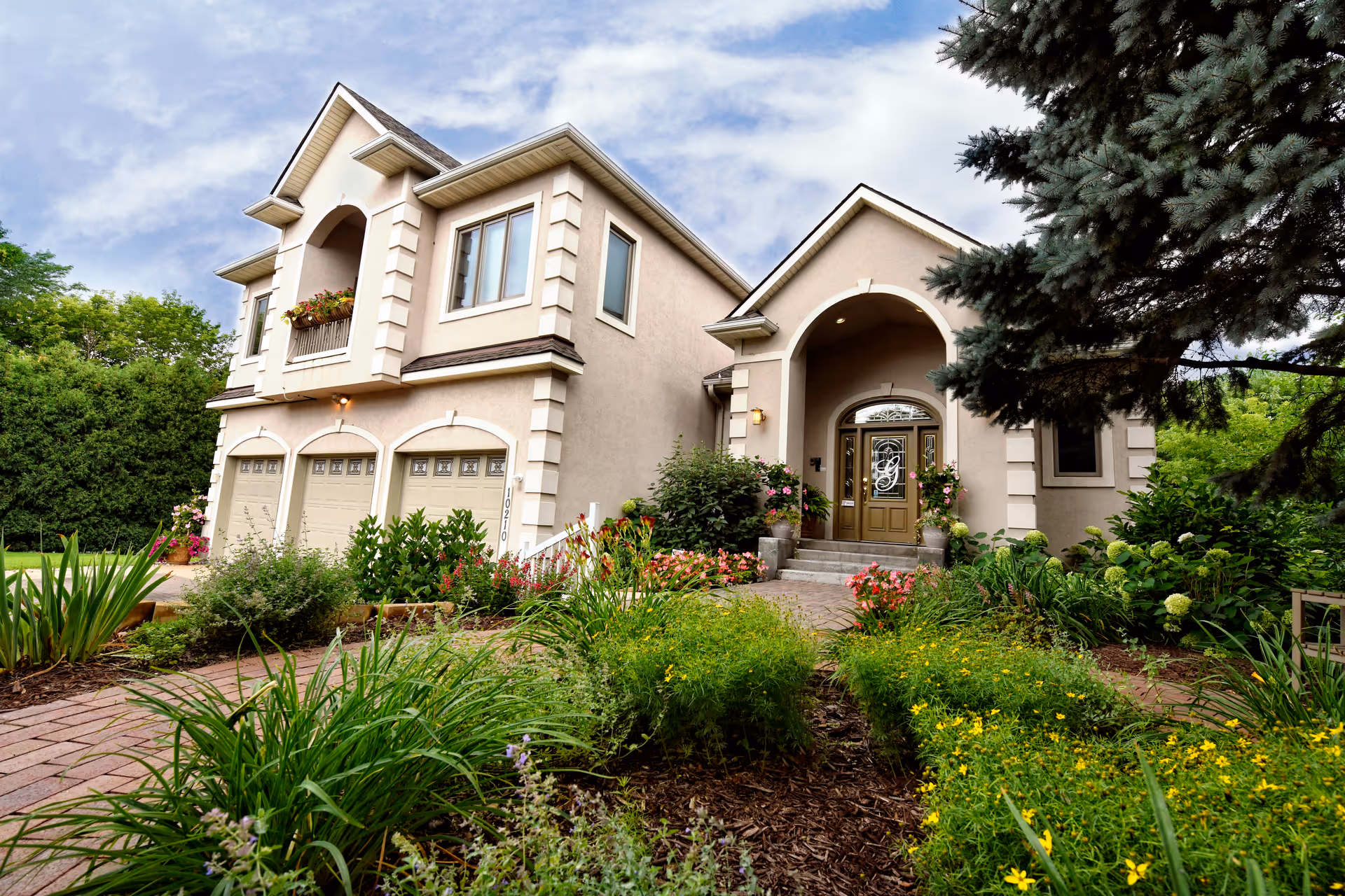 Exterior view of a large, beige two-story house with a three-car garage, arched front entrance, and well-maintained garden with various green plants and flowers in the foreground under a partly cloudy sky.