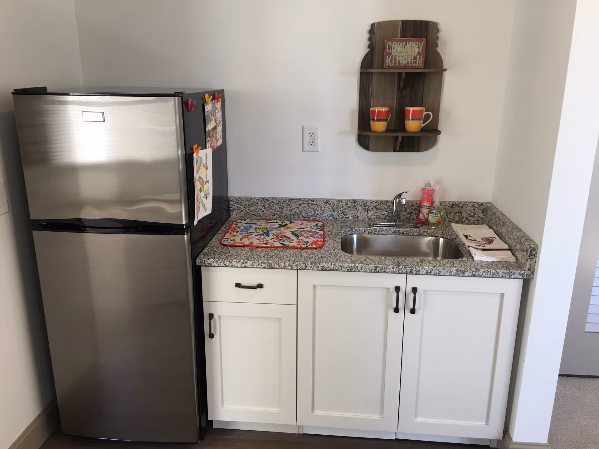 Small kitchen area with a stainless steel refrigerator on the left, white cabinets with black handles below a granite countertop, a stainless steel sink with a faucet, dish soap and a sponge on the right side of the sink, a colorful drying mat on the left side of the sink, and a small wooden wall shelf above the sink holding two colorful mugs and a sign that reads 'Country Kitchen'.