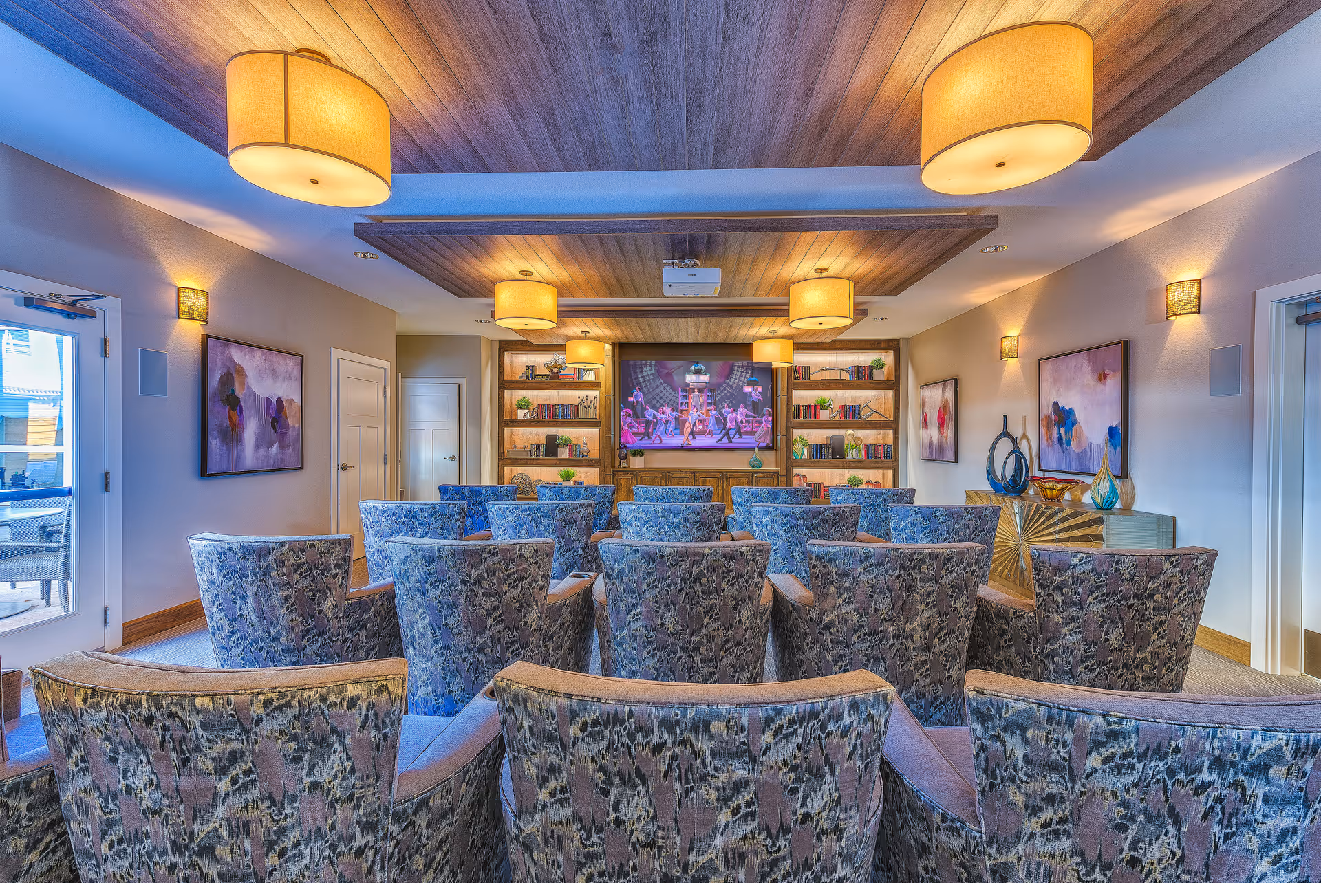 A cozy media room with multiple patterned armchairs arranged in rows facing a large flat-screen TV mounted on a wooden wall unit with shelves holding books and decorative items. The room features warm lighting from ceiling fixtures and wall sconces, abstract artwork on the walls, and a door leading to an outdoor patio area.