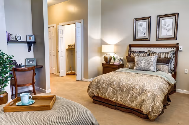 A cozy bedroom in a senior living facility featuring a neatly made bed with patterned bedding and multiple pillows. Beside the bed is a wooden nightstand with a lamp and framed photos. On the wall above the bed are two framed botanical prints. To the left, there is a small wooden desk with a chair, decorative items, and a potted plant nearby. The room has beige walls and carpeted floors, with an open doorway leading to a closet or bathroom area.