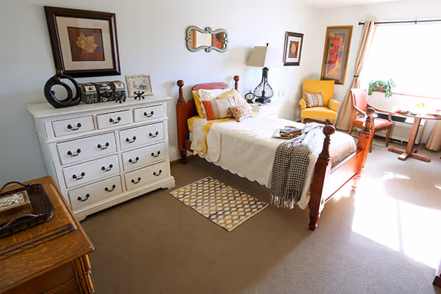 A bright and cozy bedroom with a wooden bed frame, white bedding, and multiple pillows. There is a white dresser with decorative items and framed artwork above it on the left side. A small patterned rug is placed beside the bed on a carpeted floor. Near the window, there is a yellow armchair, a wooden chair, and a small round table with a plant on it. The room is well-lit with natural light coming through the window.