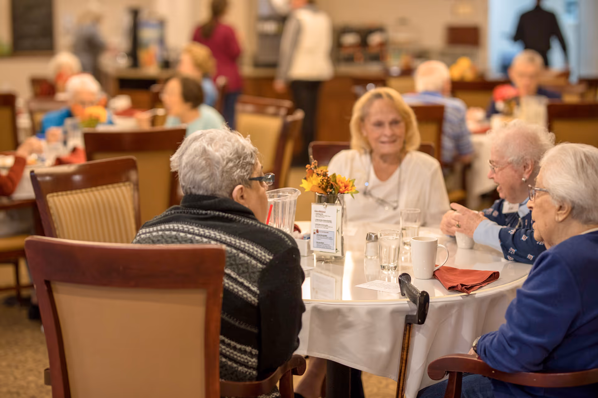 A group of elderly people sitting around a round table in a dining area, engaged in conversation. The table has glasses, cups, a small flower arrangement, and a menu or information card. Other people and tables are visible in the background.