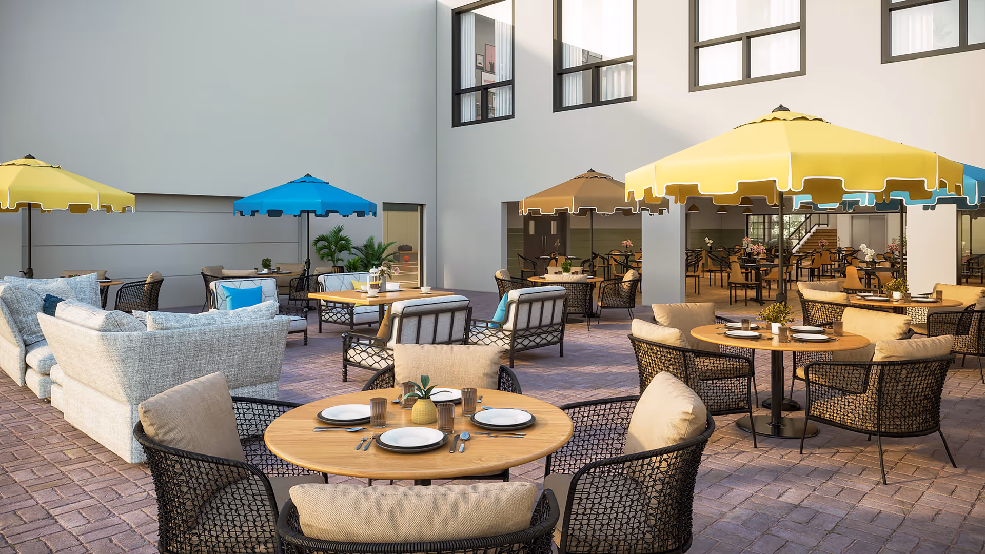 Outdoor patio area with multiple round wooden tables set with plates, glasses, and cutlery. Each table is surrounded by cushioned chairs with beige cushions. Several large umbrellas in yellow, blue, and brown provide shade. There are also cushioned sofas and armchairs arranged for seating. The area is paved with brick and is adjacent to a building with large windows.