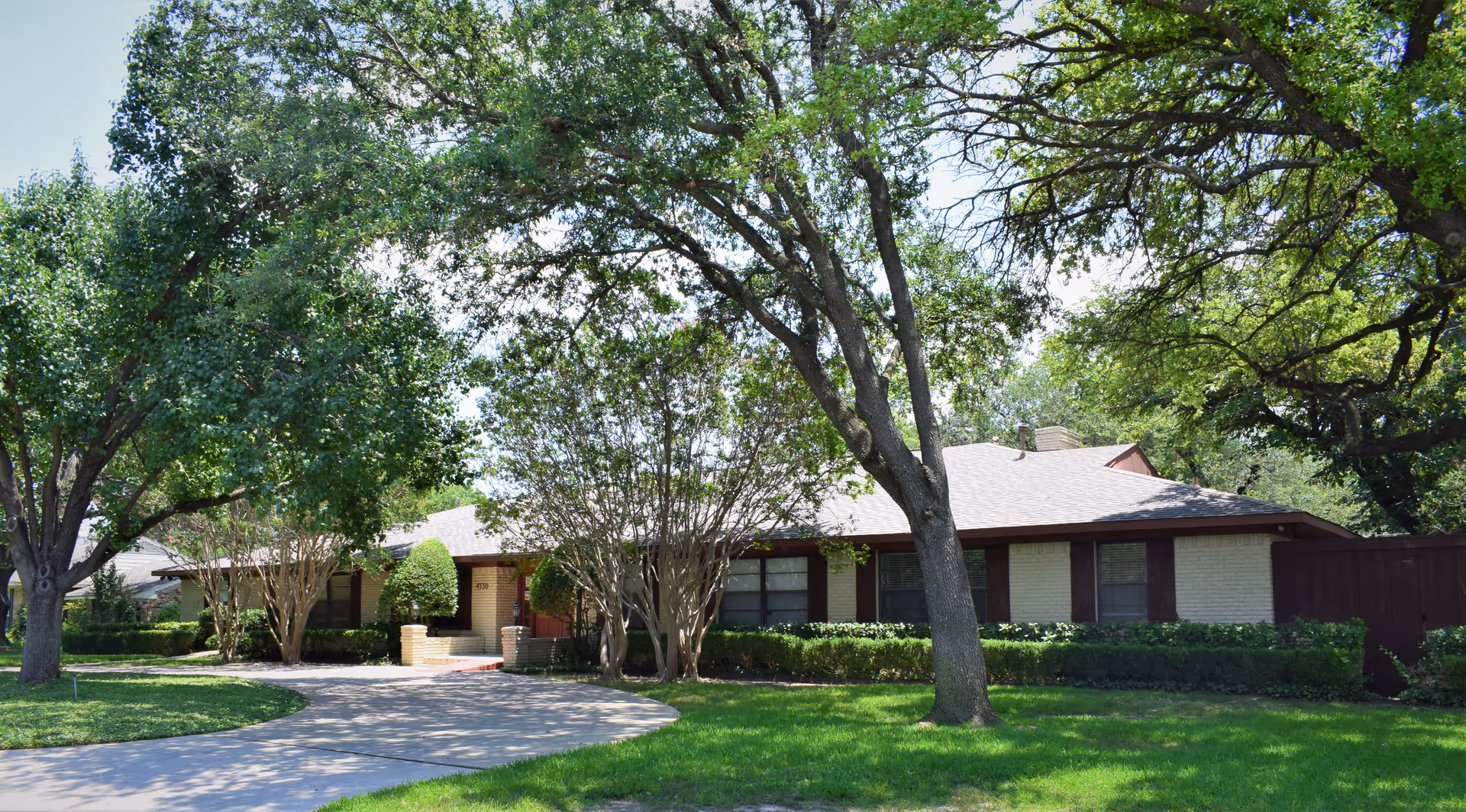 Single-story brick building with a sloped roof surrounded by large trees and green grass, featuring a curved driveway leading to the entrance.