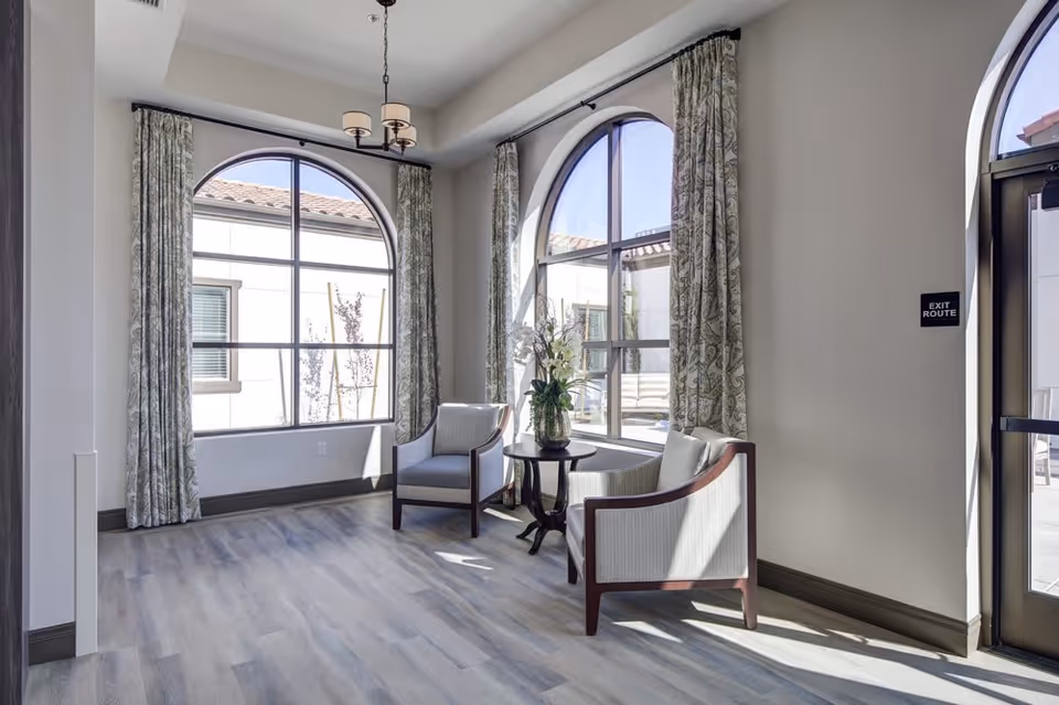 Sunlit seating area with two armchairs and a small round table in front of large arched windows with patterned curtains.
