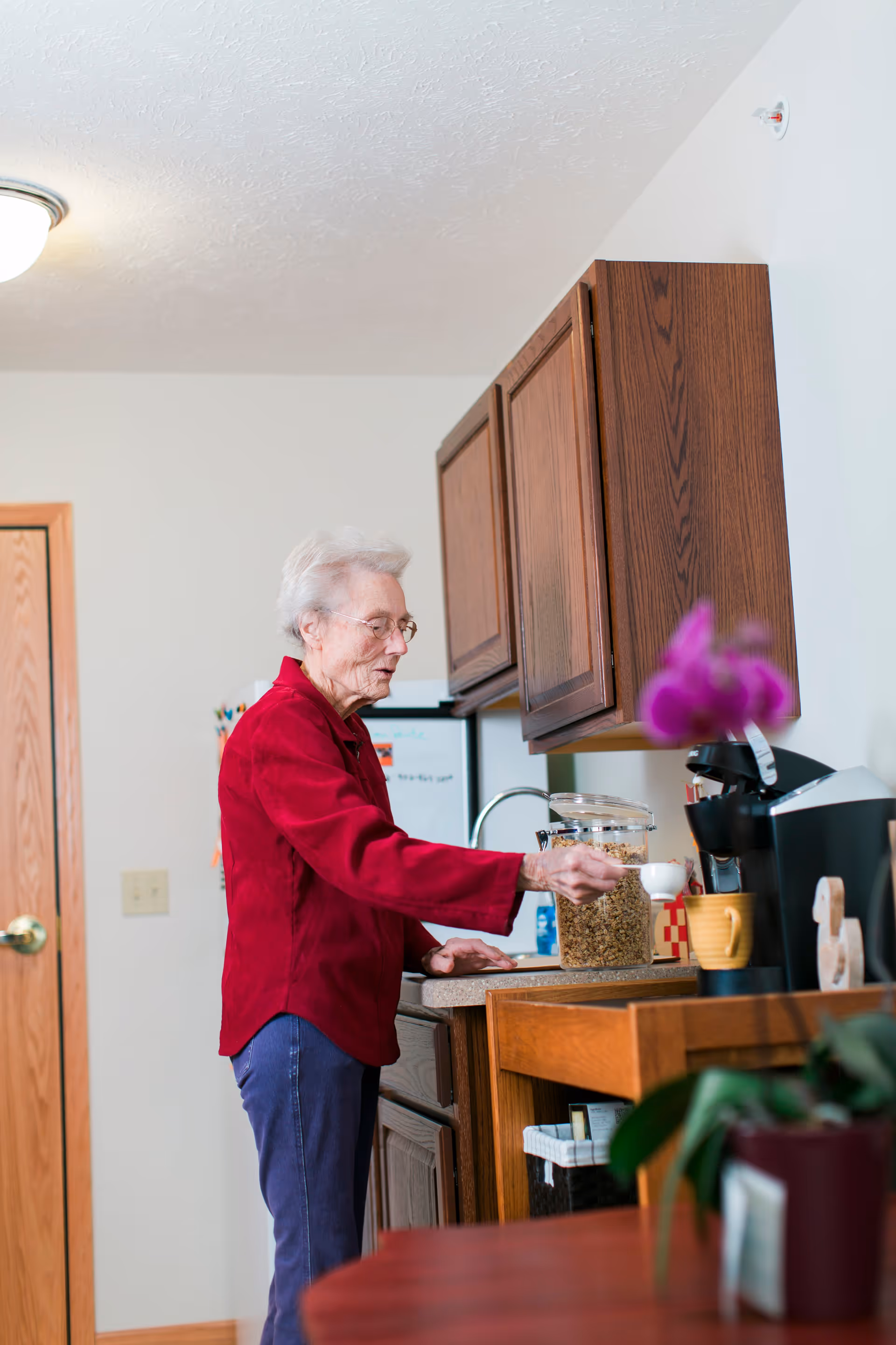 An elderly woman wearing glasses and a red sweater is standing in a kitchen area, scooping cereal from a clear container. The kitchen has wooden cabinets, a coffee maker, and a purple flower in the foreground.
