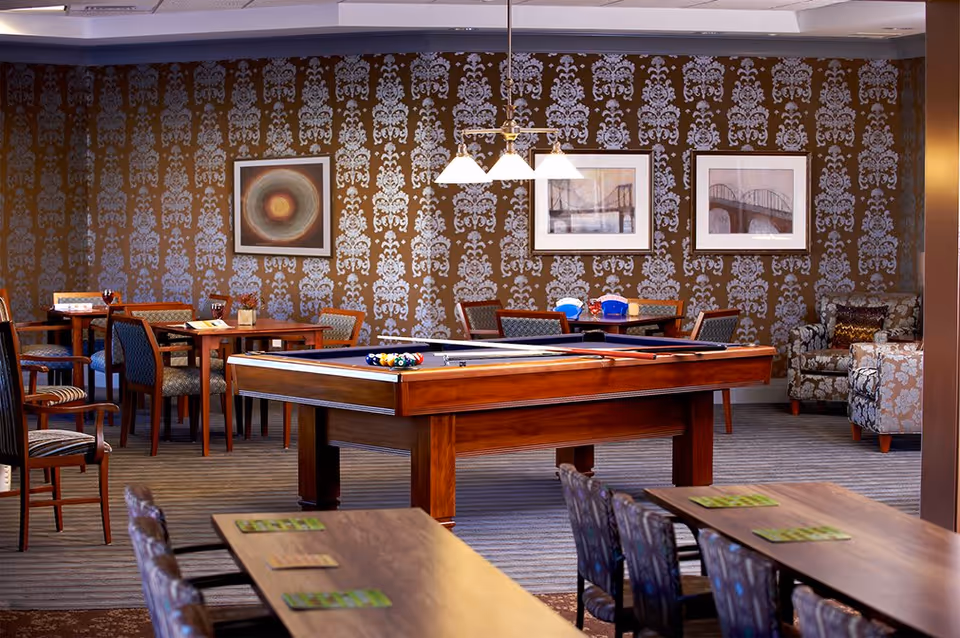 A spacious recreational room with a pool table in the center, surrounded by several tables and chairs. The walls feature ornate patterned wallpaper and framed artwork. There are comfortable armchairs in the corner and overhead lighting above the pool table.
