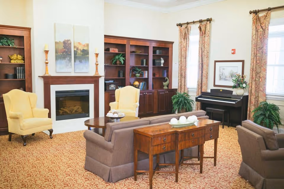 A cozy living room with two yellow armchairs, two brown sofas, a wooden coffee table, and a wooden console table with decorative items. There is a fireplace with two candles and a two-panel painting above it. Built-in wooden bookshelves filled with books and decorative items flank the fireplace. The room has patterned carpet, two windows with floral curtains, green plants, and a black upright piano with a framed picture and a vase of flowers on top.