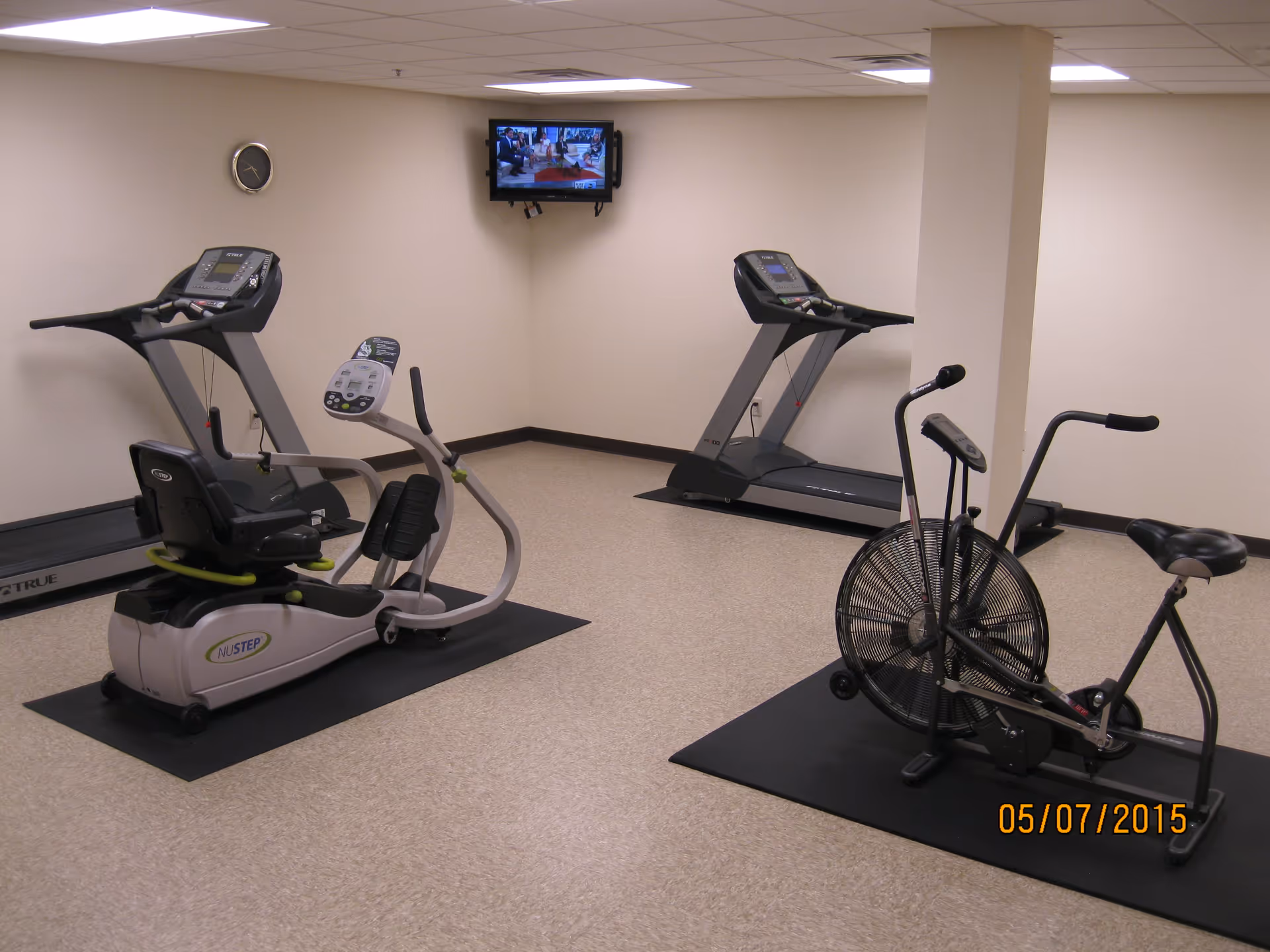 A small fitness room with two treadmills, a recumbent exercise bike, and an air resistance exercise bike. A wall clock and a small wall-mounted TV are visible on the beige walls. The floor is covered with light-colored tiles and each piece of equipment is placed on a black mat.