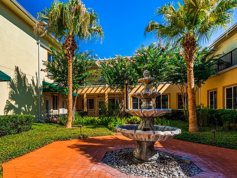 Outdoor courtyard area at The Windsor at Ortega featuring a multi-tiered stone fountain surrounded by palm trees, green shrubs, and a brick-paved walkway. The courtyard is enclosed by a two-story building with yellow walls and windows, under a clear blue sky.