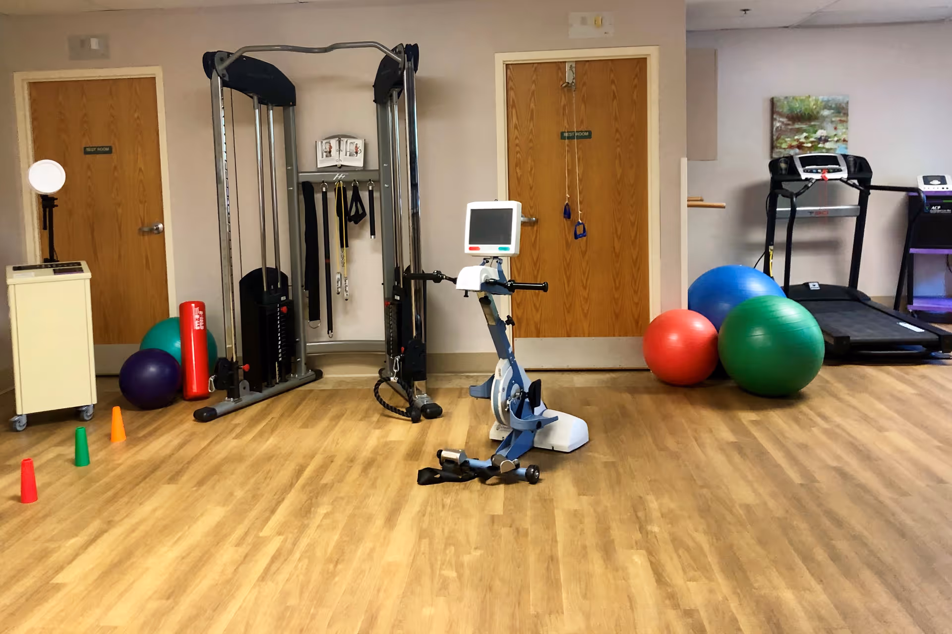 A fitness room with exercise equipment including a multi-gym machine, stationary bike with a monitor, treadmill, and various exercise balls. There are also small colored cones on the wooden floor and two closed wooden doors in the background.