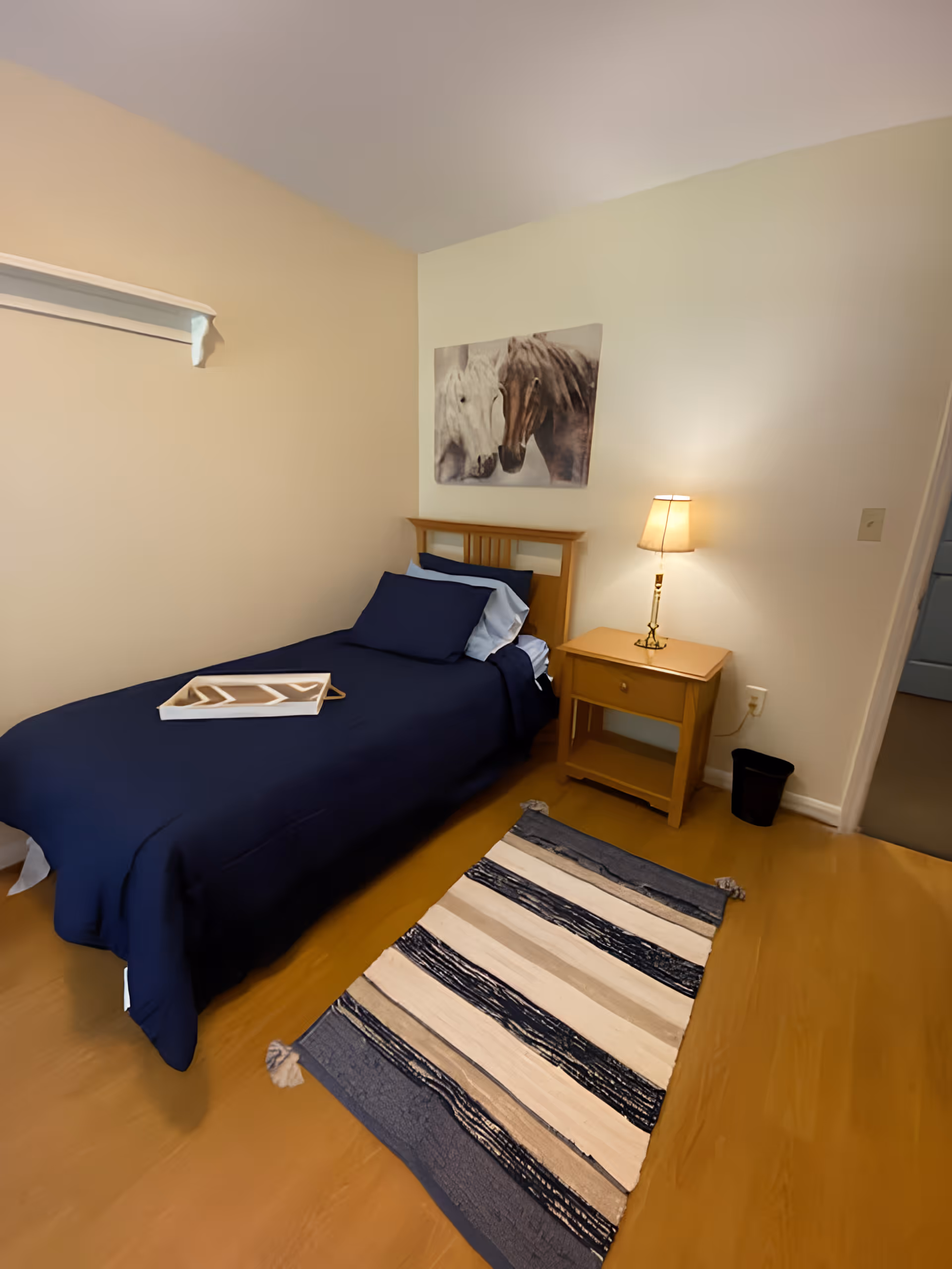 A small bedroom with a single bed covered in dark blue bedding and a white tray on top. Next to the bed is a wooden nightstand with a lit lamp. A striped rug in shades of blue, beige, and white lies on the wooden floor. Above the bed, there is a wall art featuring two horses. The walls are painted in light beige and white tones.