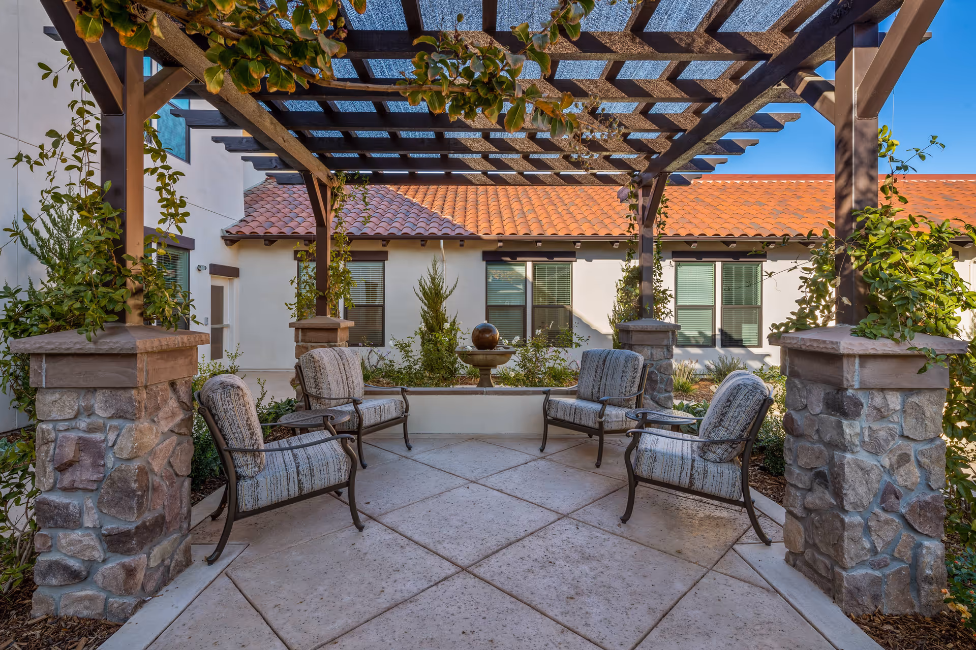 Outdoor seating area with four cushioned chairs arranged under a wooden pergola with climbing plants. The area is paved with large square tiles and surrounded by stone pillars and greenery. In the background, there is a building with a red tile roof and multiple windows.