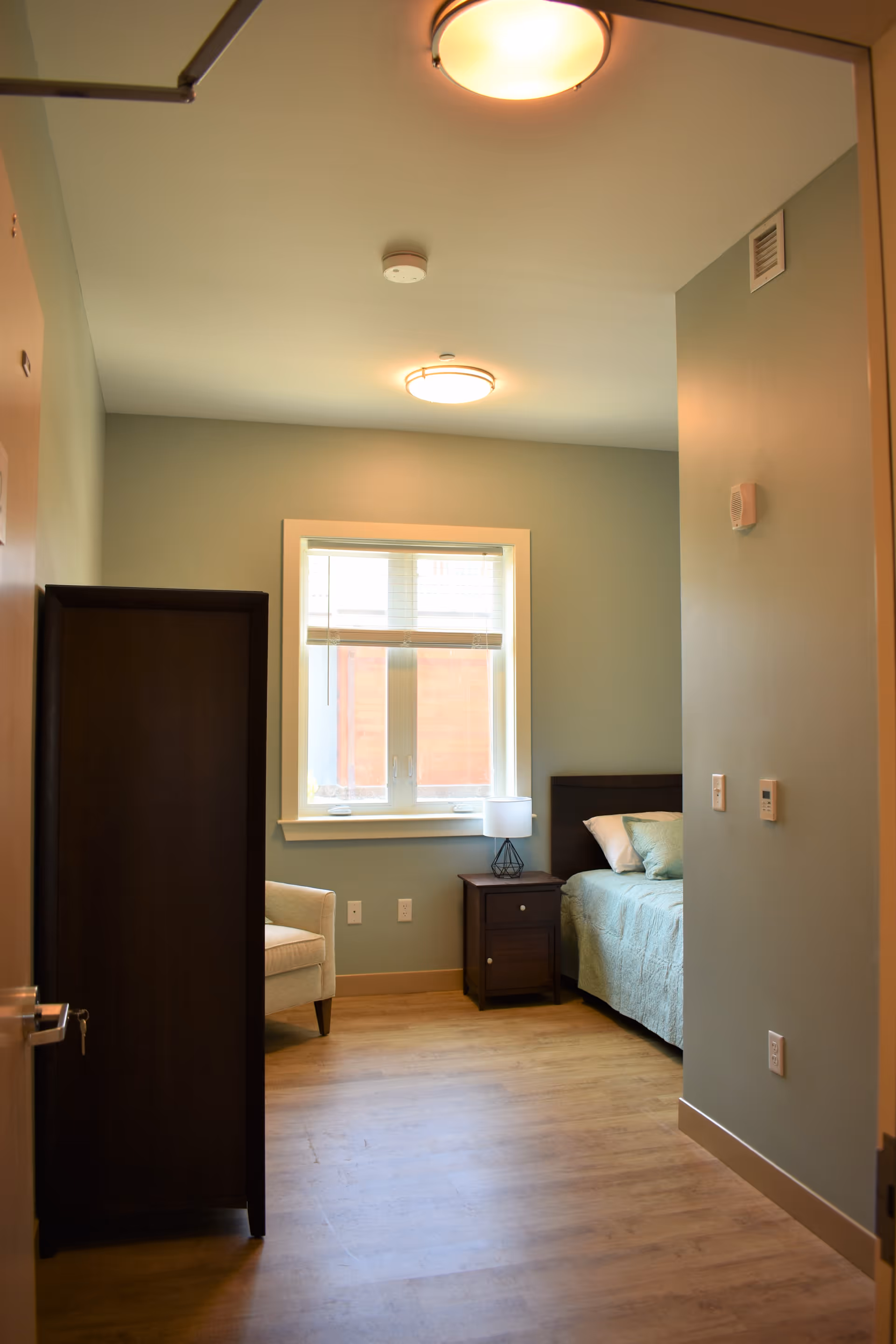 A small, cozy bedroom with light green walls and wooden flooring. The room features a single bed with a light blue bedspread and pillows, a dark wooden nightstand with a lamp, a beige armchair, and a dark wooden wardrobe. A window with white blinds lets in natural light.