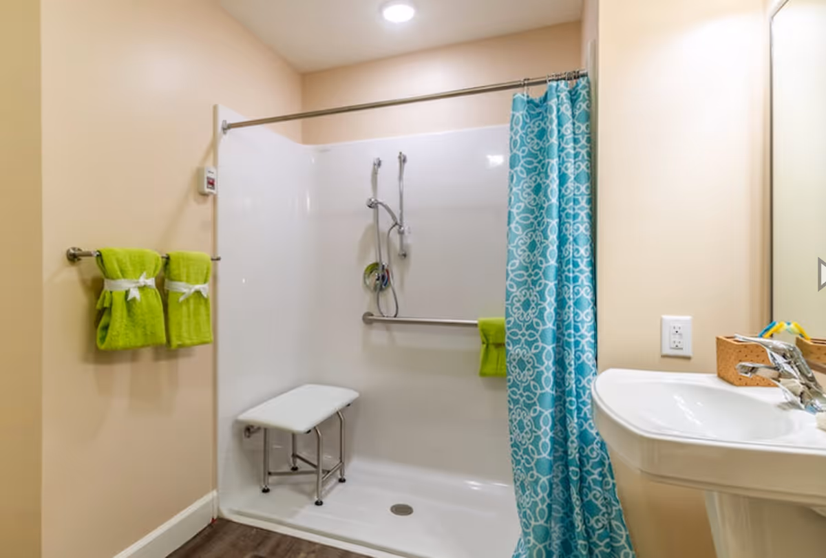 A bathroom with a walk-in shower featuring a white foldable shower seat, a handheld showerhead, and a grab bar. The shower has a blue patterned curtain partially drawn to the side. Two bright green towels are hanging on a towel rack on the left wall, and a white pedestal sink with a faucet and a small basket with toiletries is visible on the right side. The walls are painted beige and the floor has dark wood-like flooring.