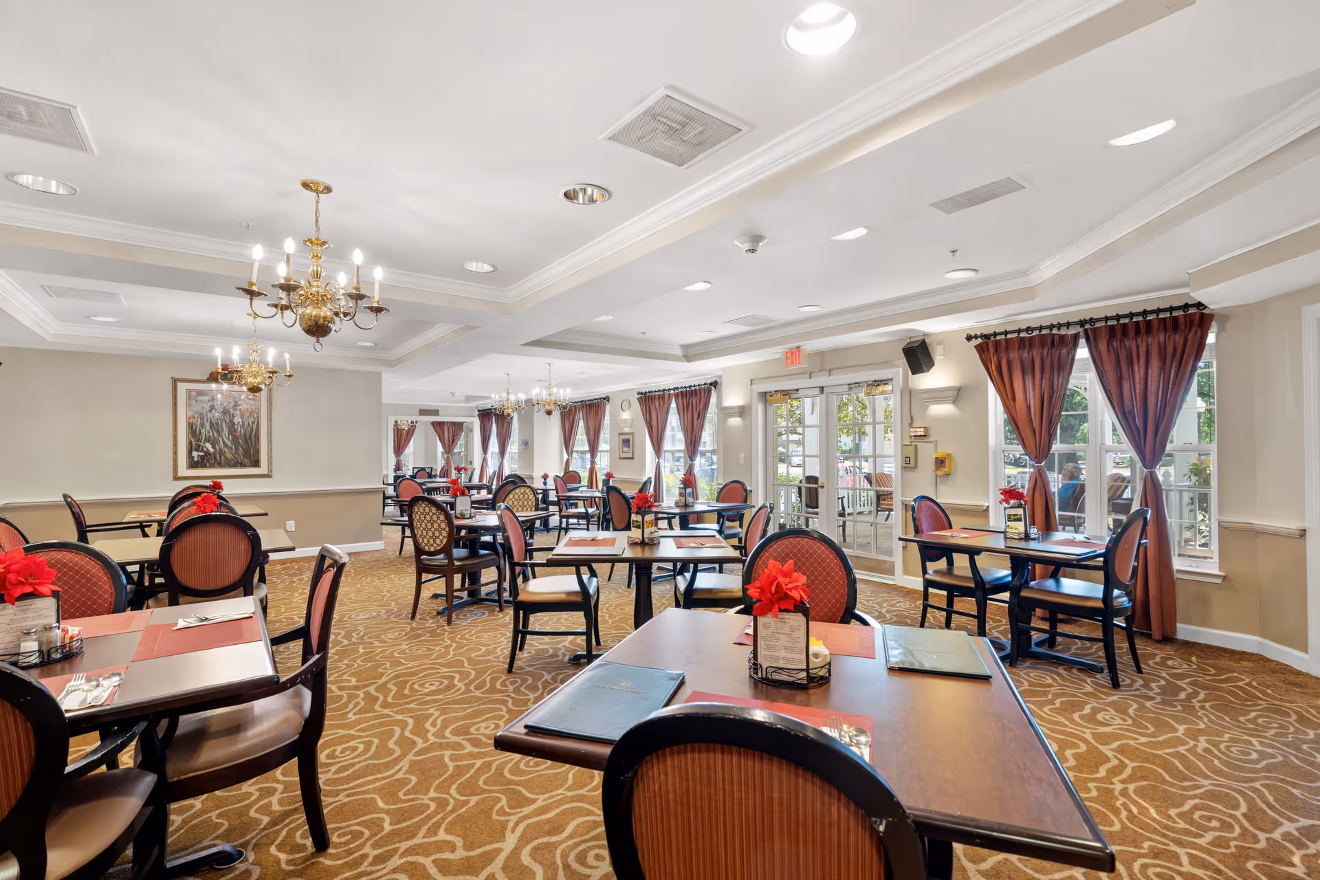 A spacious dining room with multiple tables and chairs arranged neatly. Each table has a red flower centerpiece and menus. The room features patterned carpet, chandeliers, large windows with red curtains, and glass doors leading outside.