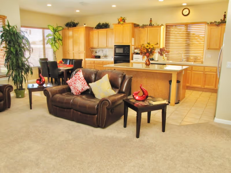 A bright and spacious open-concept living area featuring a brown leather loveseat with two decorative pillows, a small side table with a red decorative item and a book, a kitchen island with a flower vase, wooden kitchen cabinets, and a dining table with black chairs near large windows with blinds. There are also several green plants placed around the room.