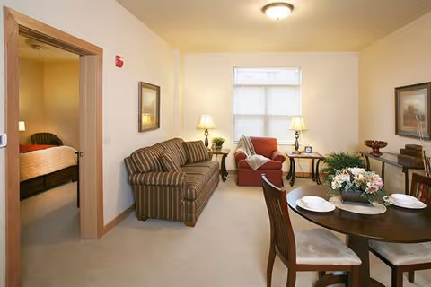 A cozy living and dining area with a striped sofa, a red armchair, two side tables with lamps, a round dining table set with plates and chairs, and a doorway leading to a bedroom with a bed and nightstand.