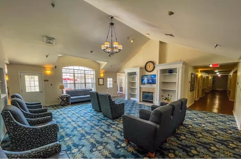 A spacious living room area in a senior living facility with patterned blue carpet, several upholstered armchairs and sofas arranged around a fireplace with built-in shelves. A large clock is mounted above the fireplace, and a chandelier hangs from the vaulted ceiling. There is a large window letting in natural light and a hallway leading to other rooms.