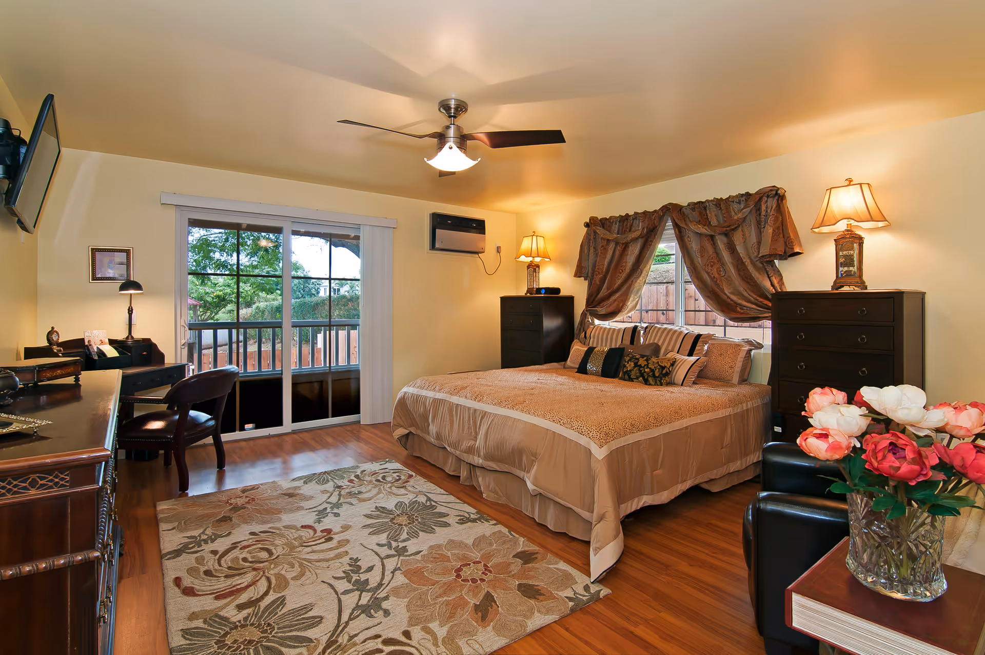 A cozy bedroom with a large bed covered in beige bedding and multiple pillows. The room features wooden flooring, a floral area rug, two dark wooden dressers with lamps on top, a window with decorative curtains, a sliding glass door leading outside, a ceiling fan with light, a desk with a chair, and a vase of flowers on a side table.