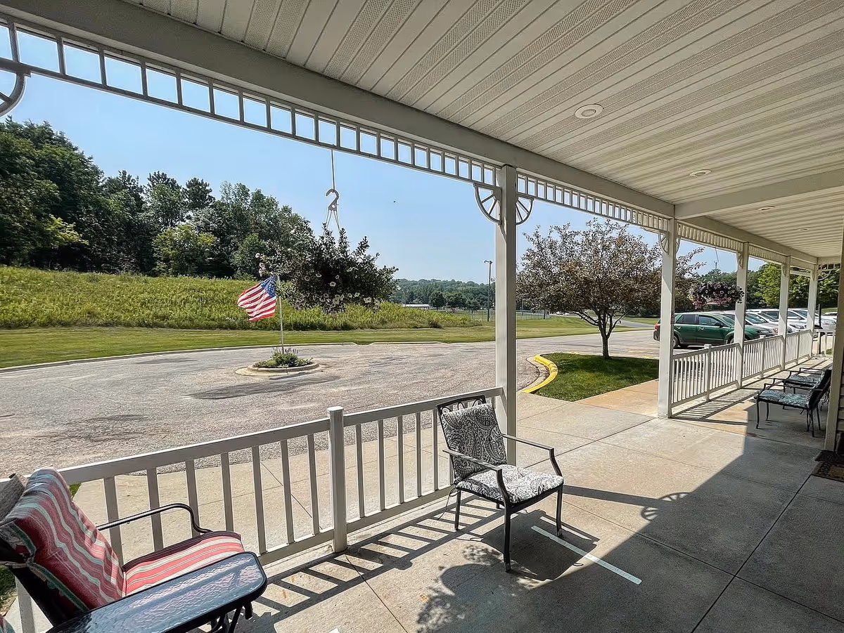 Covered outdoor patio area with several cushioned chairs arranged along a white railing. Hanging flower baskets are suspended from the patio roof. Beyond the patio is a circular driveway with an American flag planted in the center, surrounded by greenery and trees under a clear blue sky.