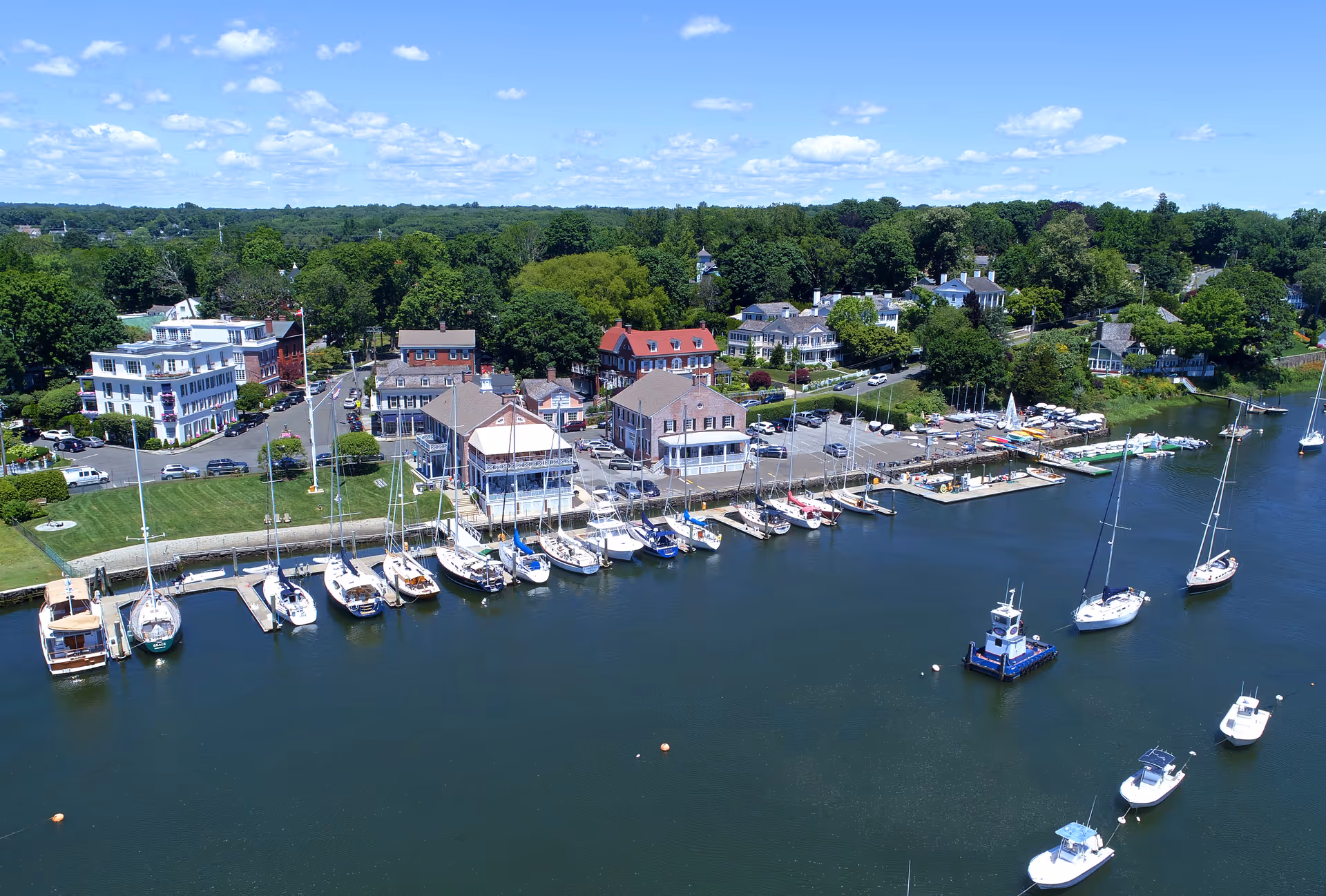 Aerial view of a waterfront area with multiple sailboats and small boats docked along piers. Several buildings, including houses and commercial structures, are visible near the shoreline, surrounded by green trees and a clear blue sky with scattered clouds.