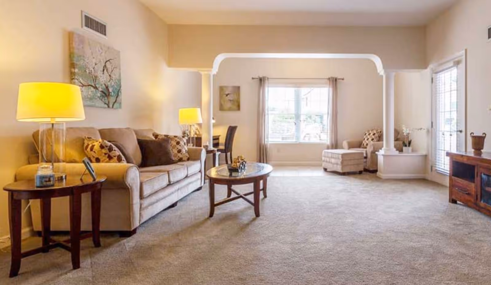 A spacious living room with beige walls and carpet. The room features a beige sofa with patterned cushions, two side tables with lamps, a round wooden coffee table, a single armchair with an ottoman near a large window with curtains, and a wooden media console. The room is well-lit with natural light coming through the window and glass door.