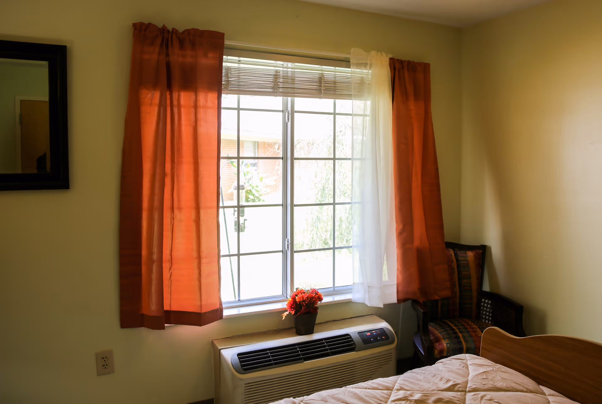 A small bedroom with a window covered by orange and white curtains. Below the window is an air conditioning unit with a small potted plant on top. To the right of the window is a wooden chair with a colorful cushion. Part of a bed with a beige comforter is visible in the foreground.