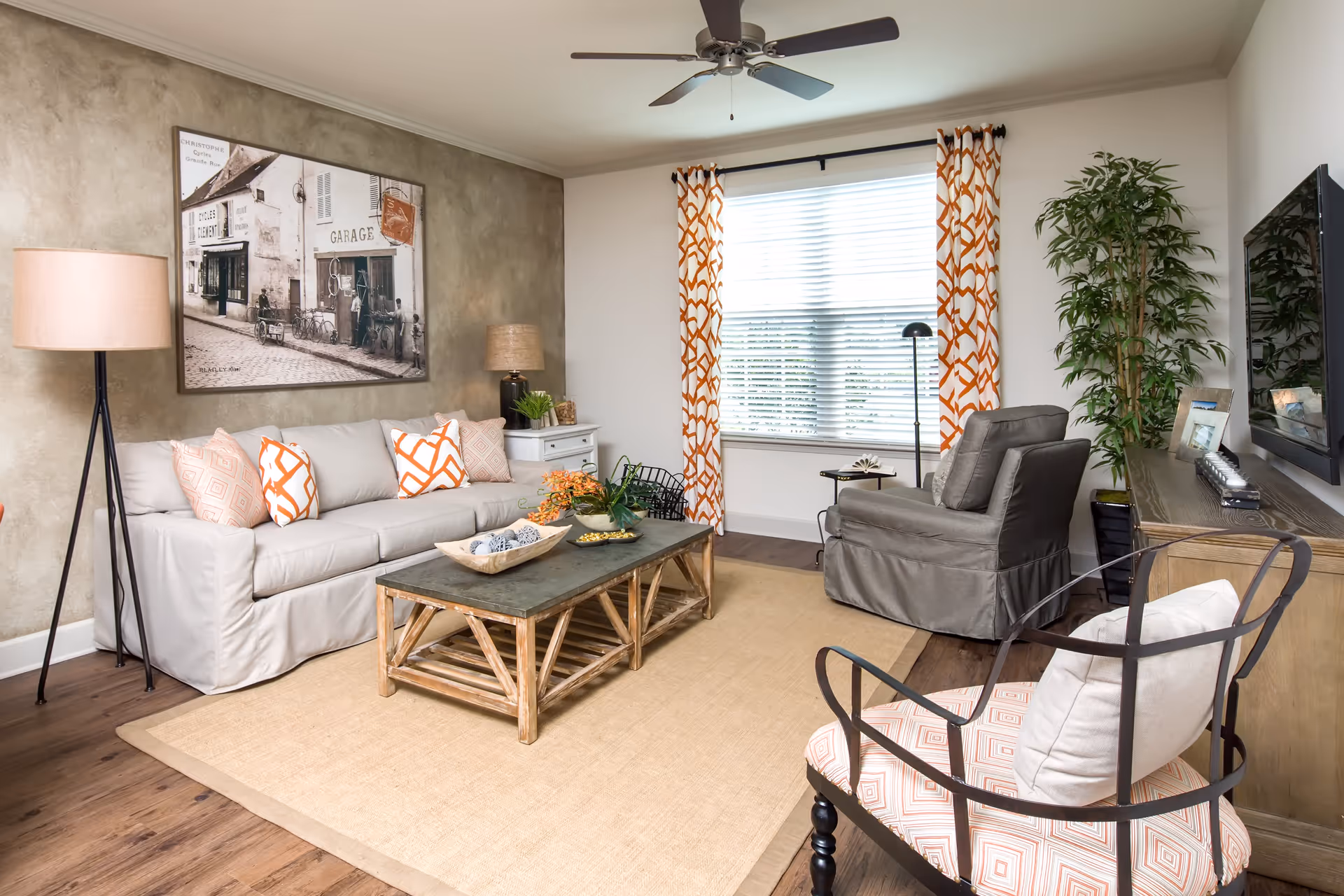 A cozy living room with a beige sofa adorned with patterned orange and white pillows, a wooden coffee table with decorative items, a gray armchair, and a cushioned chair with a geometric pattern. The room features a large window with orange and white curtains, a floor lamp, a side table with a lamp and plants, a large wall-mounted TV, and a framed vintage-style photograph on the wall.