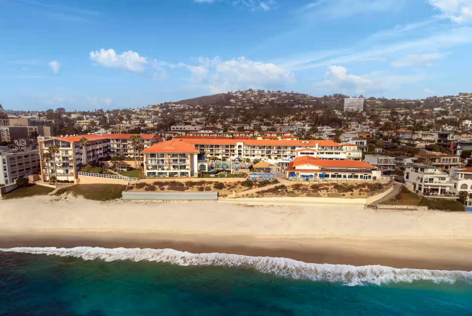 Aerial view of White Sands La Jolla facility located along a sandy beach with clear blue ocean water in the foreground. The building has multiple stories with red-tiled roofs and balconies facing the ocean. Behind the facility, there is a hillside with numerous houses and buildings under a partly cloudy blue sky.