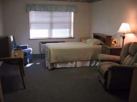 A bedroom in a senior living facility with a bed, sofa, chair, and dresser.