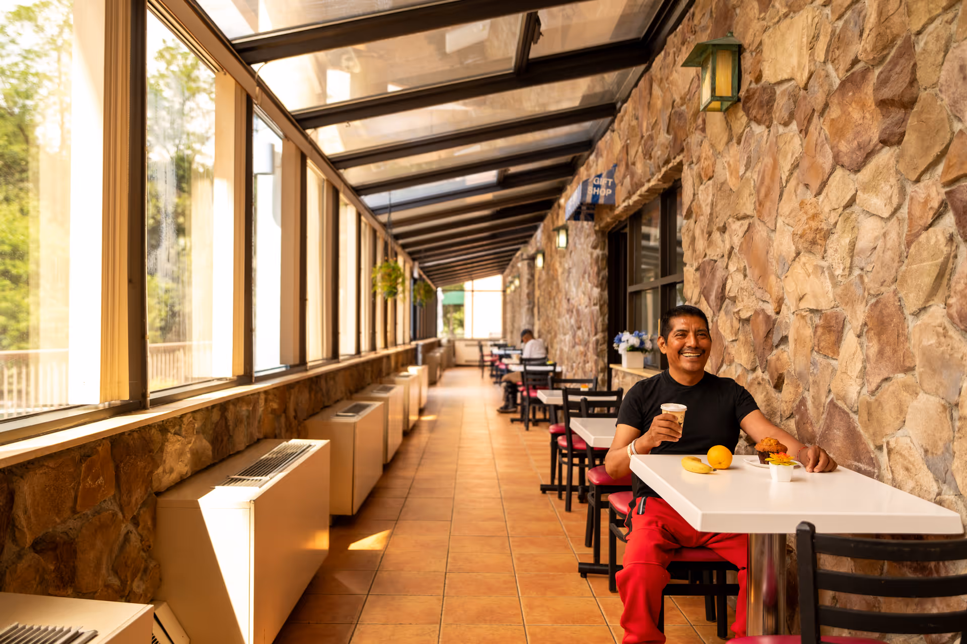 A man sitting at a white table in a sunlit indoor corridor with large windows on one side and a stone wall on the other. He is smiling and holding a cup, with food items including a banana, an orange, and a muffin on the table. Several other tables and chairs line the corridor, and a sign for a gift shop is visible on the stone wall.