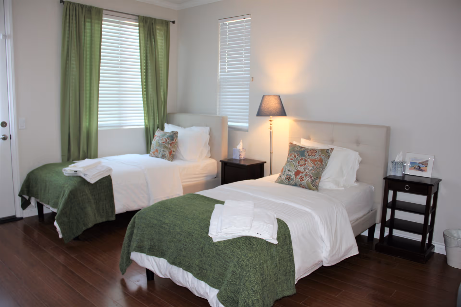 A bright bedroom with two single beds, each made with white linens, green blankets, and decorative pillows. Between the beds are two dark wooden nightstands, one with a lamp and tissue box, and the other with a tissue box and framed photo. The room has wooden flooring, two windows with white blinds and green curtains, and a white wall.